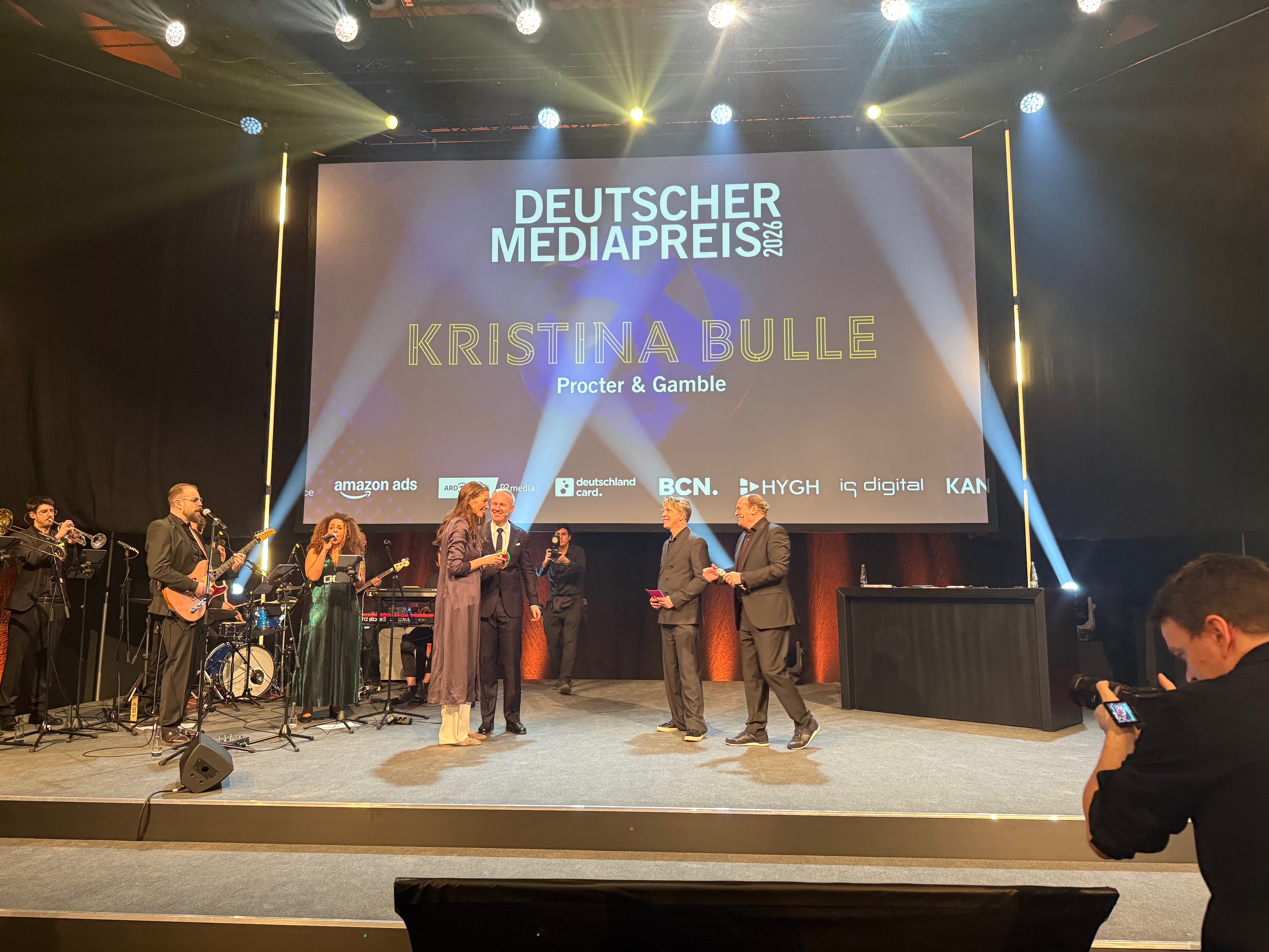 Kristina Bulle is on stage and being handed the award. There is a band in the background and a phottographer is picturing the moment.